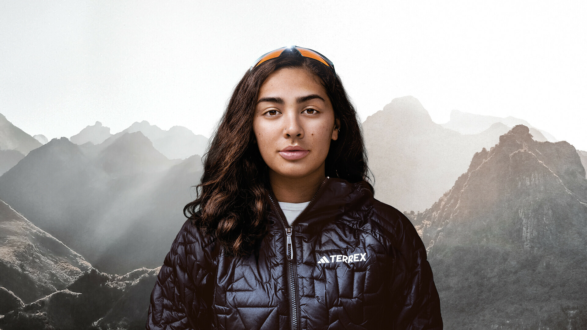 Portrait de Samantha Soriano regardant directement la caméra. Elle porte une veste d'extérieur noire et des lunettes de soleil sur la tête. En arrière-plan, on voit des sommets de montagnes embrumés.