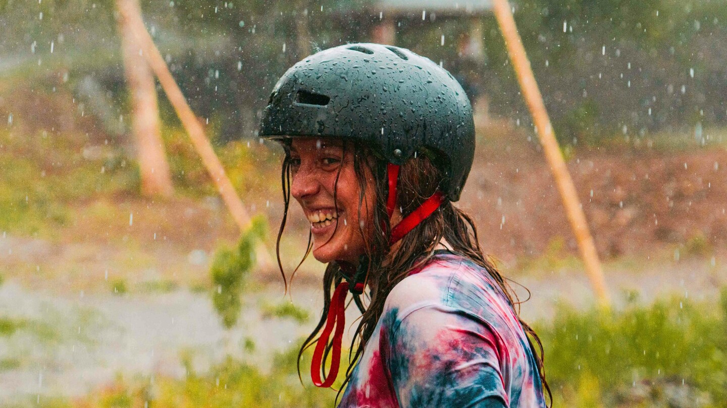 Anna Pixner, une skateboardeuse de descente, sourit sous la pluie en portant un casque noir et un T-shirt coloré tie-dye. À l'arrière-plan, on aperçoit un cadre rural avec un pavillon et plusieurs personnes.