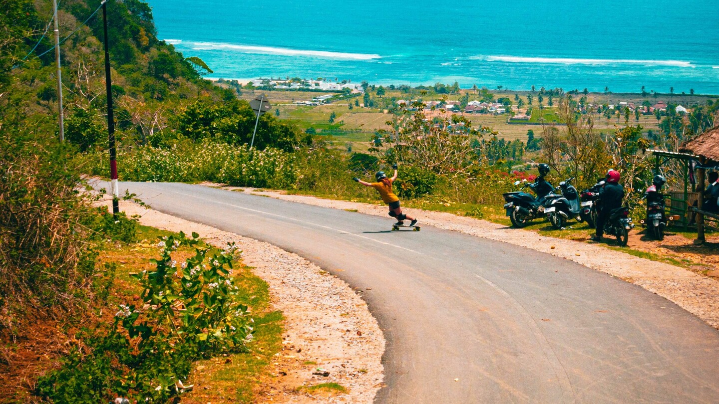 Une skateboardeuse de descente dévale une route sinueuse menant à une vue côtière magnifique avec une mer turquoise. La route serpente à travers un paysage verdoyant, bordée de végétation et de quelques motos sur le bord de la route. En arrière-plan, l'océan s'étend sous un ciel bleu éclatant avec des nuages blancs, apportant une atmosphère estivale et aventureuse à la scène.