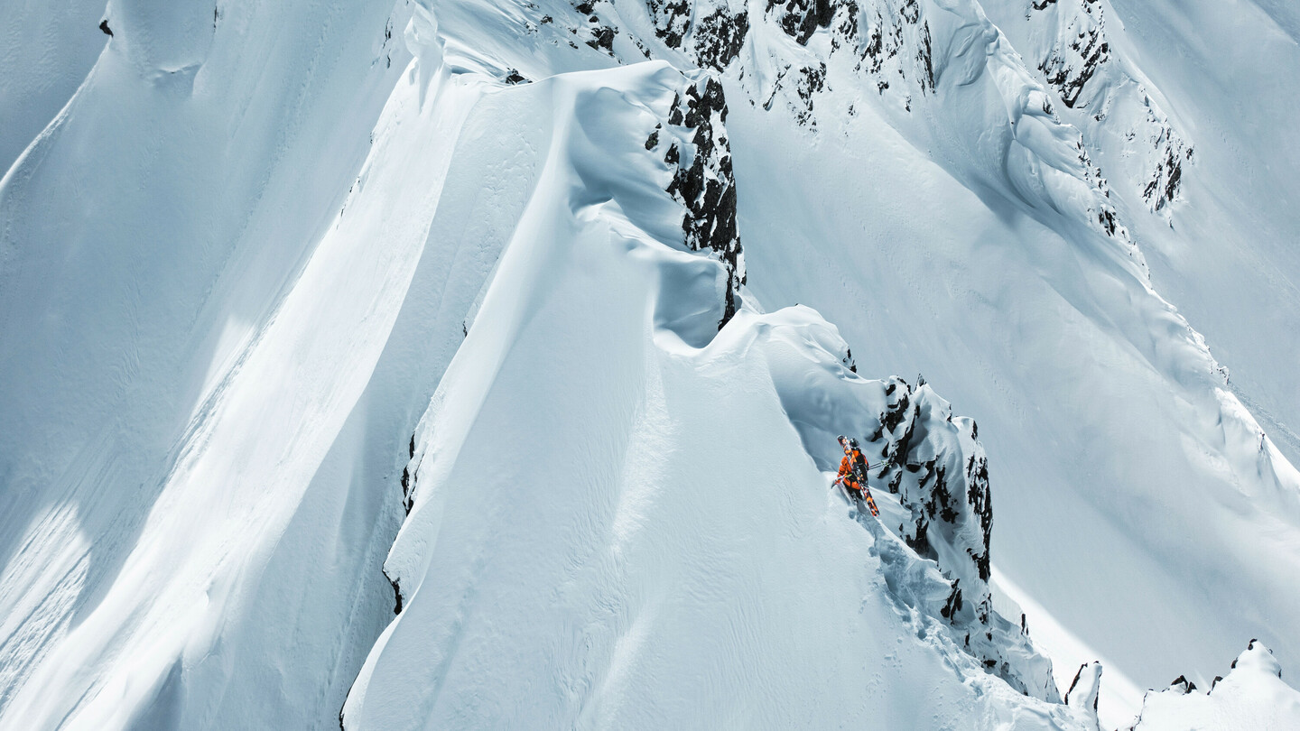 Skieur en veste orange naviguant avec précaution le long d'une crête enneigée et étroite dans un paysage de montagne dramatique.