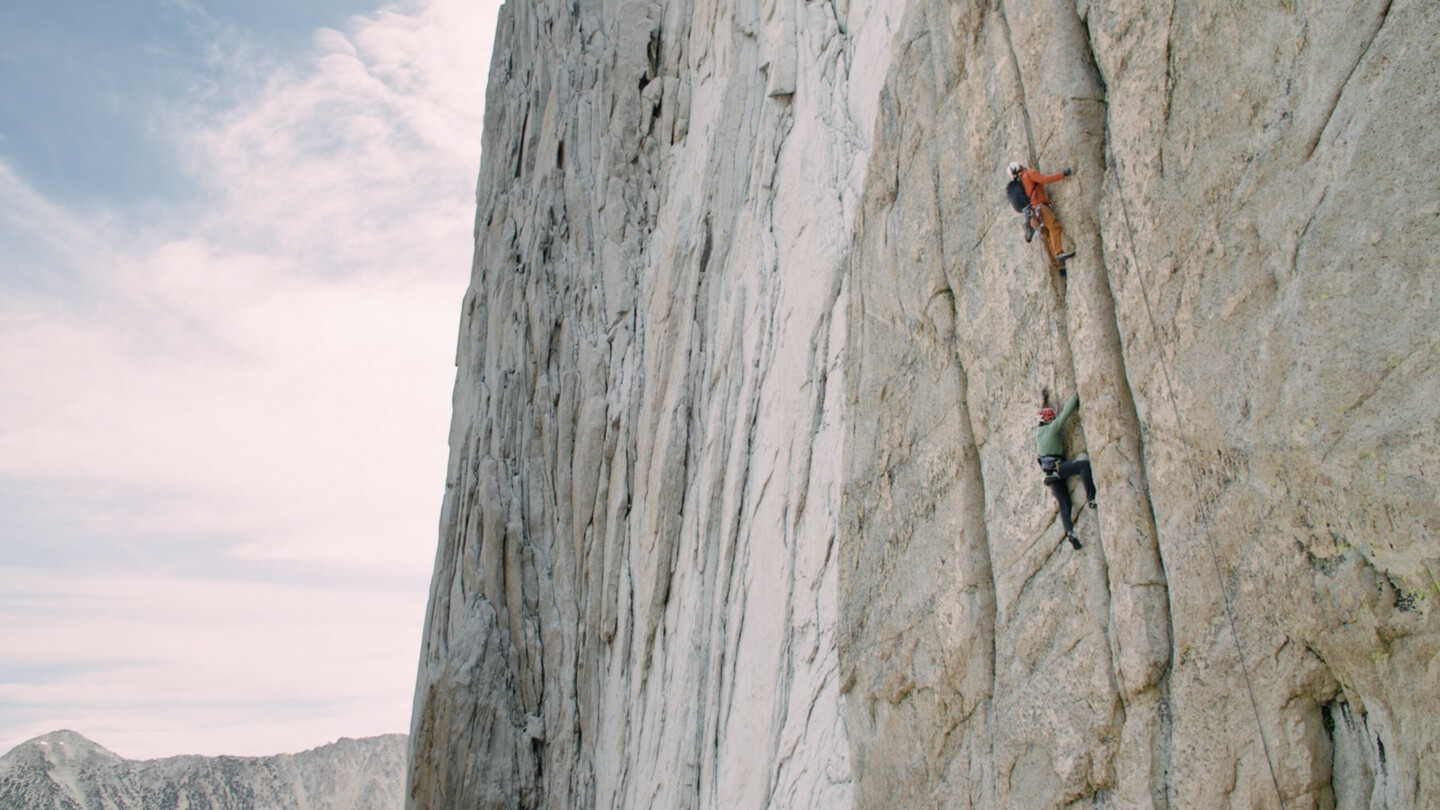 Deux grimpeurs en équipement d'escalade avec des casques escaladent une paroi rocheuse raide et verticale. Le grimpeur du haut porte une veste orange, tandis que celui du bas porte une veste verte. La paroi massive s'élève haut dans le ciel, sur fond de ciel clair et légèrement nuageux.