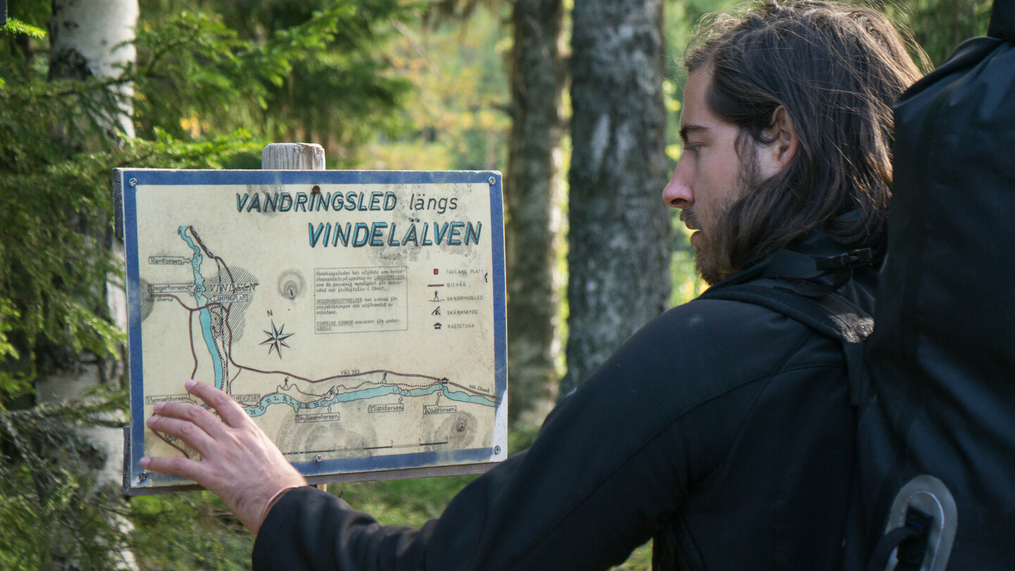 A man with shoulder-length hair and a large backpack examines a hiking map labeled “Vandringsled längs Vindelälven” in a forest. He is touching the map with his hand and appears focused. Trees and dense woodland surround him.