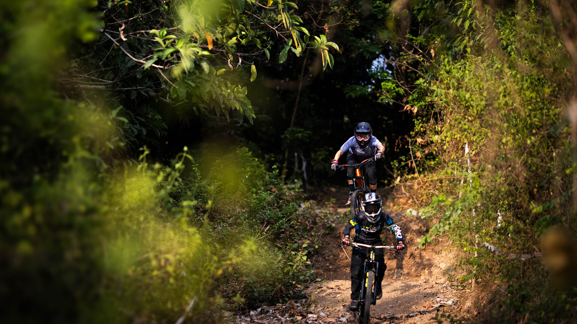 Zwei Mountainbiker fahren einen schmalen, von B&auml;umen umgebenen Pfad hinunter. Der vordere Fahrer tr&auml;gt eine schwarze Ausr&uuml;stung mit einem Helm, w&auml;hrend der hintere Fahrer eine graue Kleidung und eine Schutzbrille tr&auml;gt.