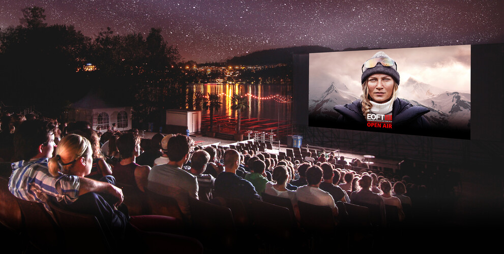 Ein Outdoor-Kino am See mit einem grossen Publikum, das unter dem Sternenhimmel einen Film auf einer Leinwand anschaut. Auf der Leinwand ist eine Frau mit Outdoor-Kleidung und Berglandschaft im Hintergrund zu sehen, darüber steht „EOFT OPEN AIR“.